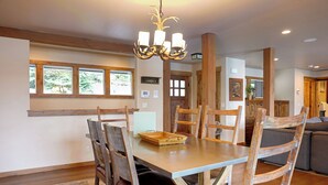 A dining area with a wooden table and chairs, a chandelier, and a view through large windows. - Kokanee Bay Beach House Lake Coeur d'Alene! (Coeur d'Alene)