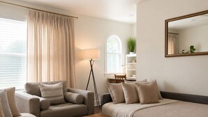 Ein modernes Hotelzimmer mit einer Sofagarnitur, einem Holztisch, einer Stehlampe, einem Fenster mit Vorhängen, einer Pflanze und einem Spiegel an der Wand.