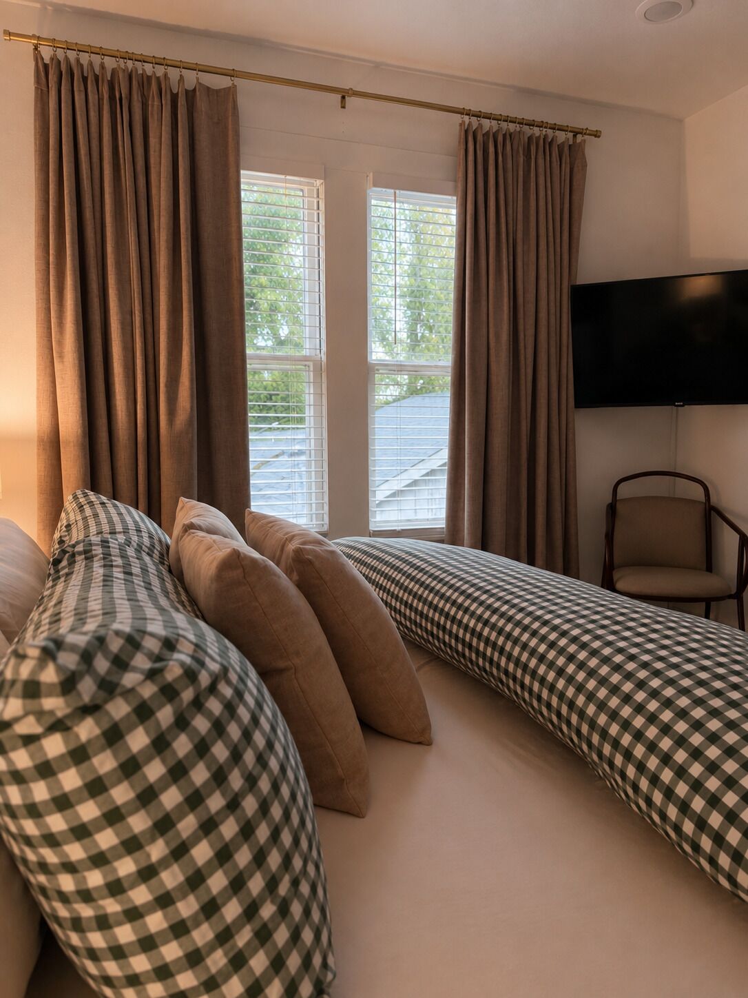 Een slaapkamer met een bed, kussens, een stoel, een televisie en een raam met gordijnen.