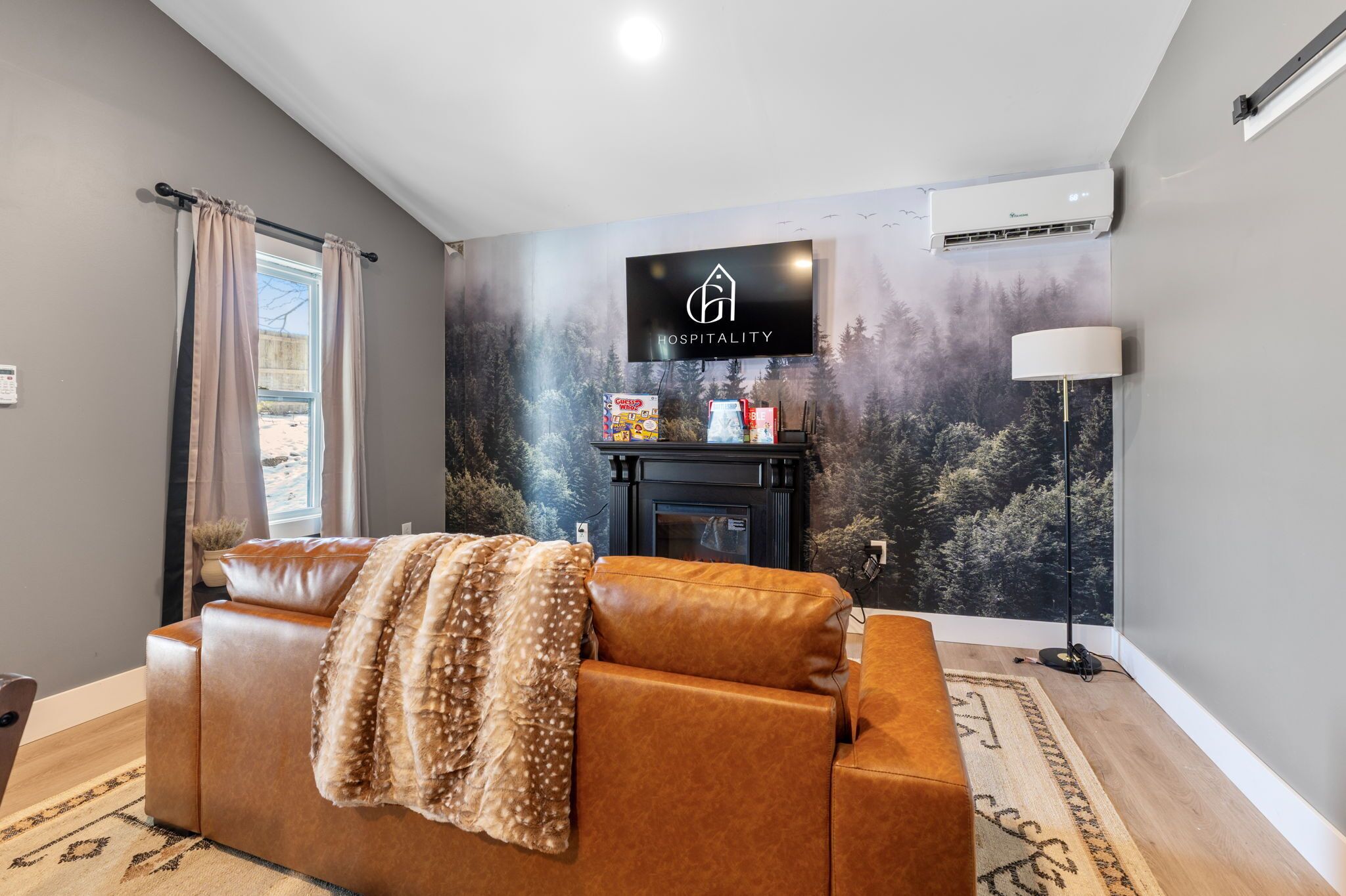 A modern living room with a brown leather sofa, a fireplace, a mounted TV displaying a 'Hospitality' logo, and a forest-themed wall mural.