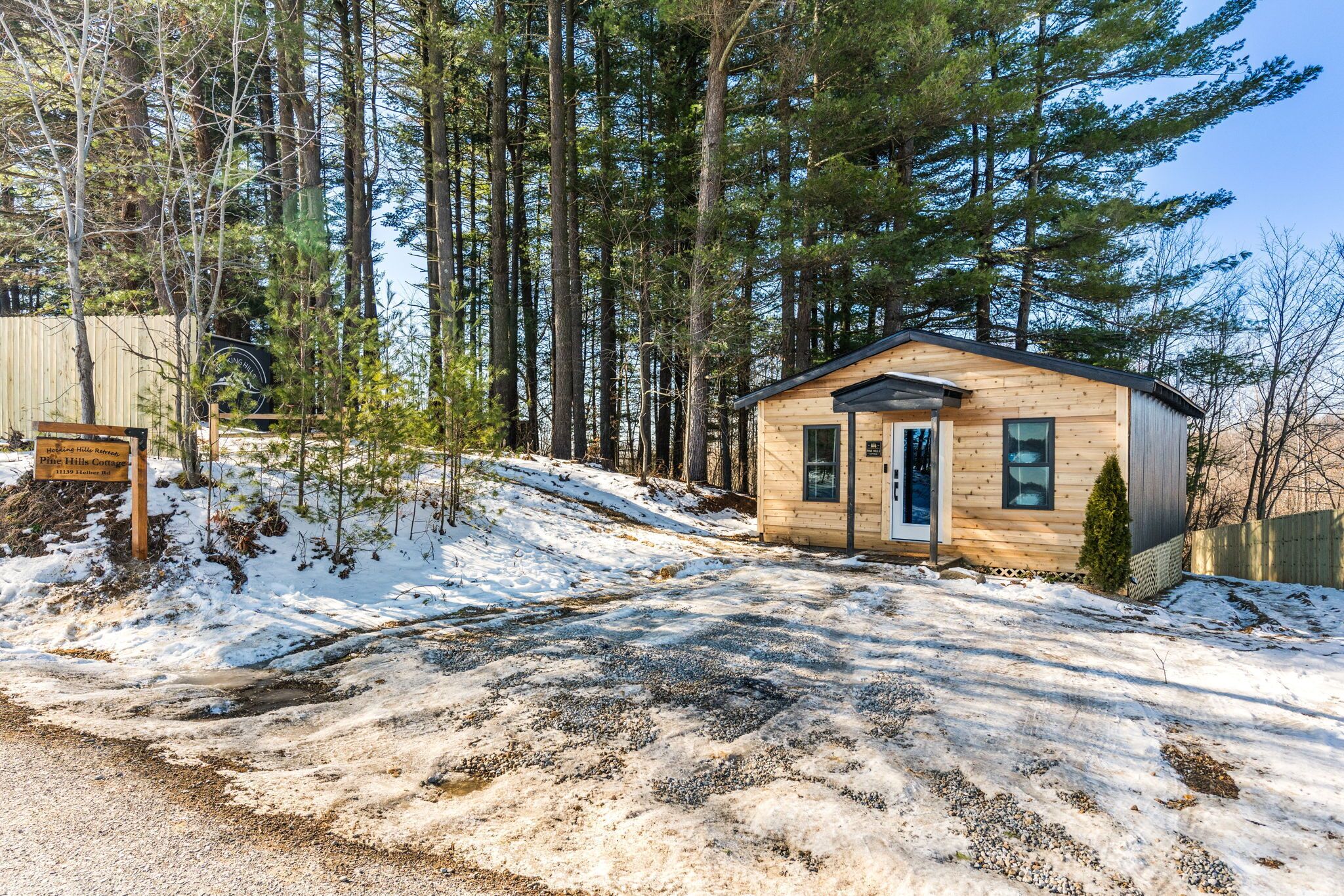 Une petite cabane en bois, dotée d’une porte et de fenêtres, entourée de grands arbres et bordée d’un sol enneigé.