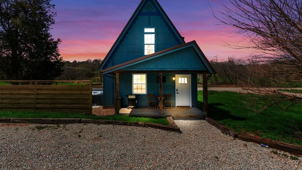 Uma casa azul com telhado triangular, varanda com duas cadeiras e um caminho de acesso de cascalho.