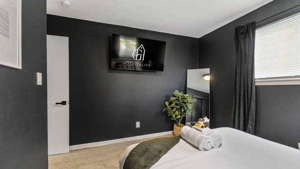 Habitación de hotel moderna con una pared negra, un televisor de pantalla plana, una planta en maceta, una cama con ropa de cama blanca y una ventana con cortinas.