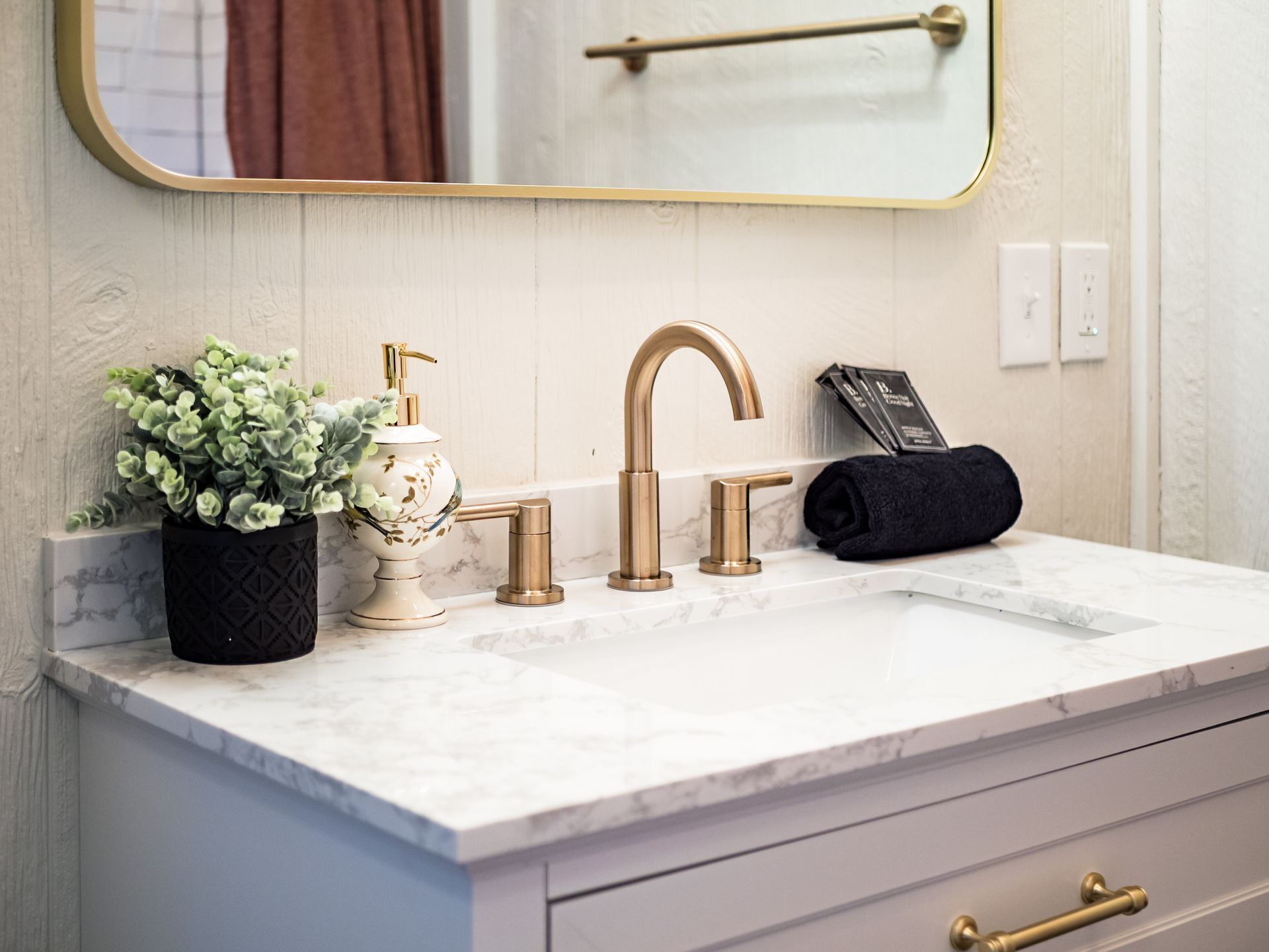 Une salle de bain avec un comptoir en marbre, un robinet doré, un lavabo sur pied blanc, une serviette noire et une plante en pot.
