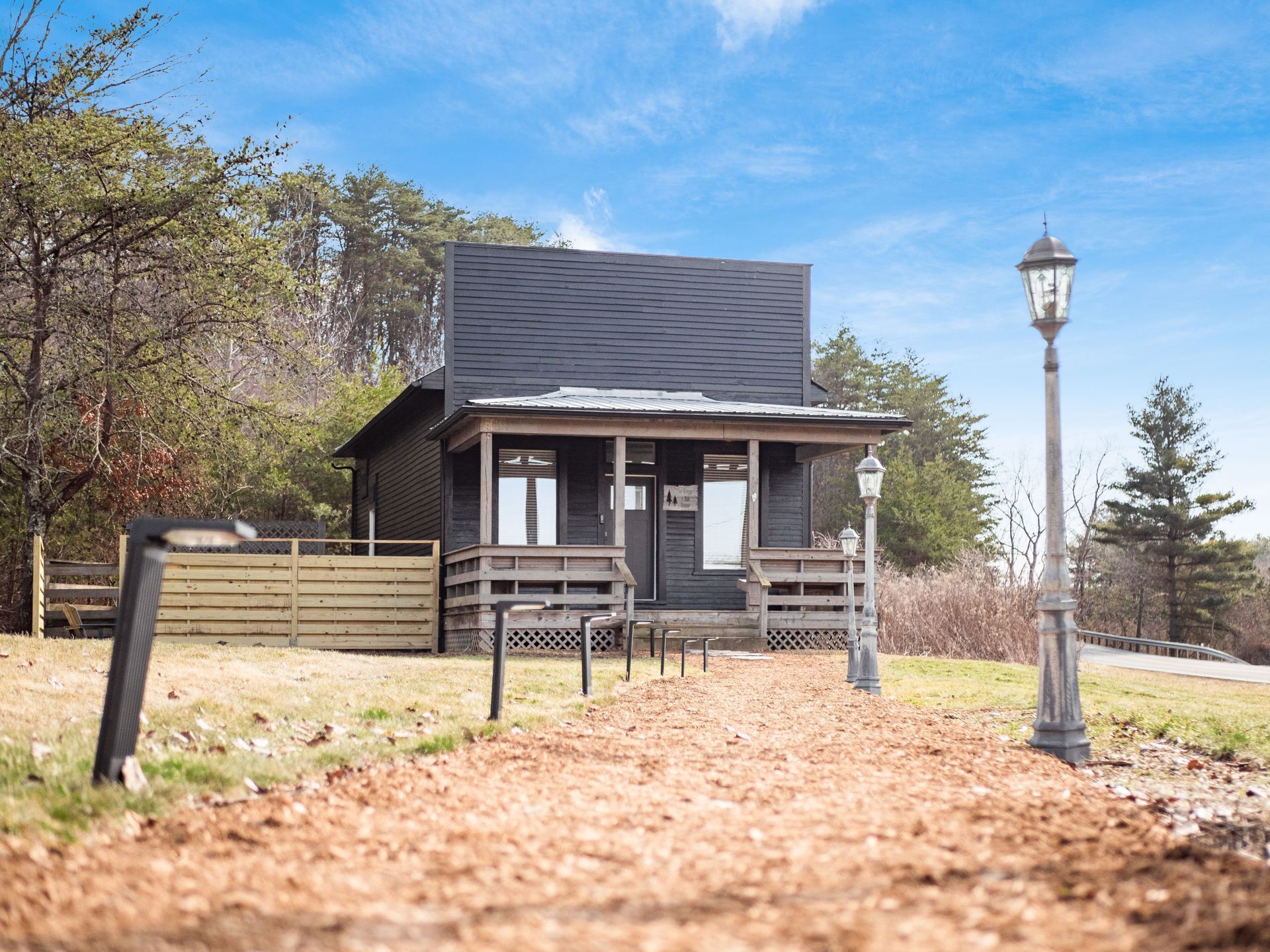 A small, single-story building with a porch and a lamp post, surrounded by trees and a dirt path.