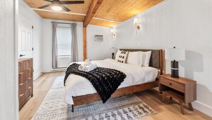 Un dormitorio con techo de madera, una cama con ropa blanca, una mesita de noche y un velador.
