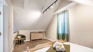 Quarto com teto inclinado, cama com manta verde, mesinha de cabeceira com um vaso e uma cadeira verde.