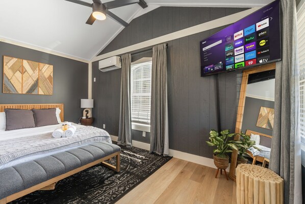 A modern hotel room with a large bed, a flat-screen TV mounted on the wall, a bedside table with a lamp, and a potted plant.