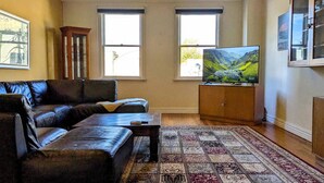 A living room with a leather sofa, a coffee table, a patterned rug, a television on a wooden cabinet, and two windows with blinds. - Heart of Surrey Hills (Parking On Request) (Surrey Hills)