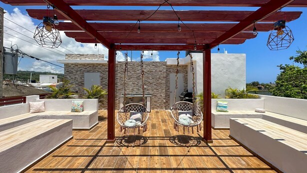 A covered patio area with two wicker chairs and a table, wooden flooring, and a stone wall in the background. - Cape Coral 1 (Cabrera)
