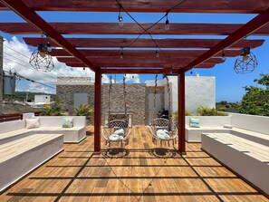 A covered patio area with two wicker chairs and a table, wooden flooring, and a stone wall in the background. - Cape Coral 1 (Cabrera)