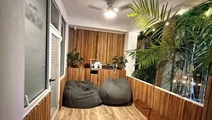 Una habitación acogedora con suelo de madera, ventilador de techo y ventana con cortinas. Hay dos pufs y una pequeña mesa con altavoces.