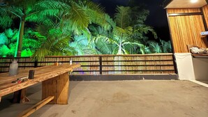 Un área de estar al aire libre con un banco y una mesa de madera, rodeada por una cerca de bambú y palmeras.