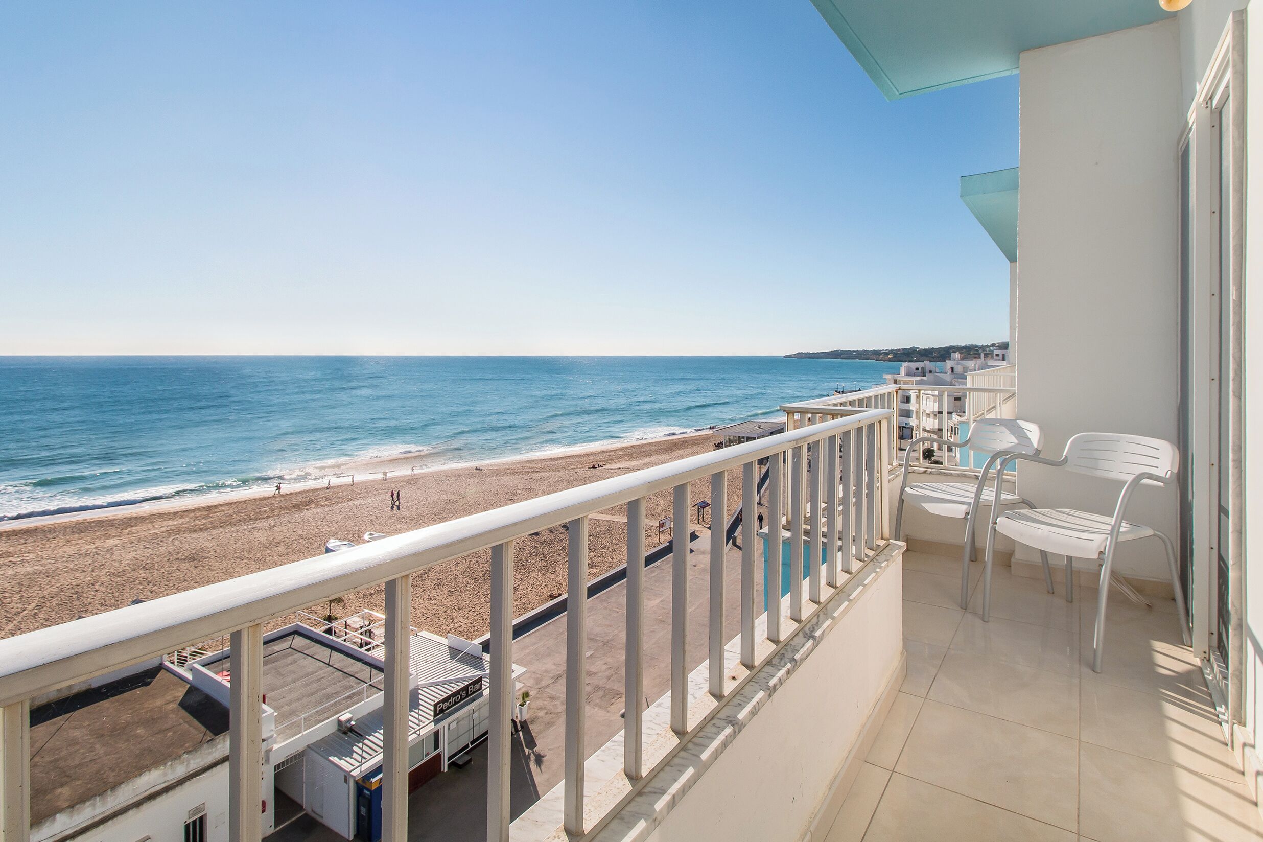 Un balcó amb vista a la platja i l'oceà. Hi ha cadires blanques i una taula al balcó.