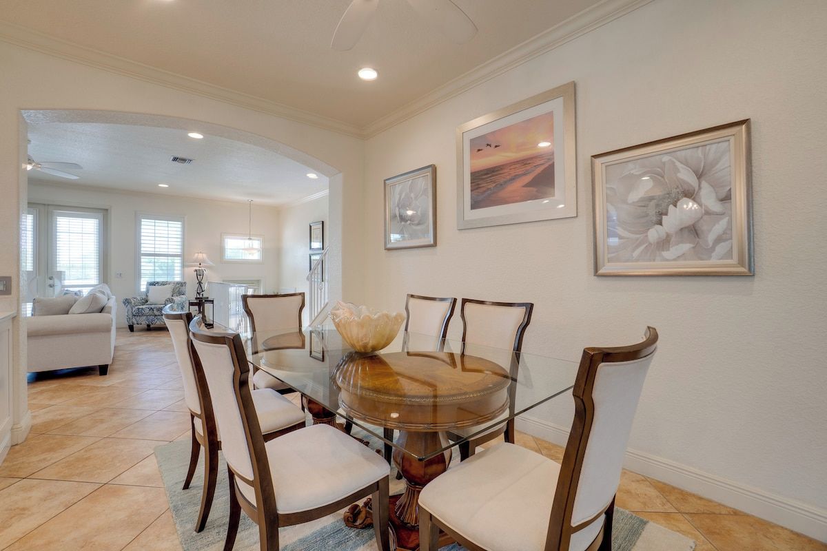 A dining area with a glass-top table surrounded by chairs, framed artwork on the walls, and a view into the adjacent living room.