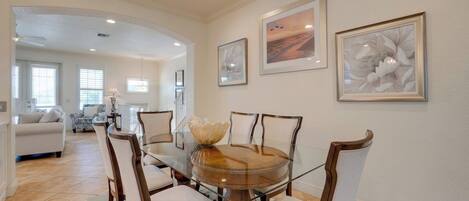A dining area with a glass-top table surrounded by chairs, framed artwork on the walls, and a view into the adjacent living room.