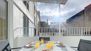 Ein Esstisch, gedeckt für das Frühstück, mit Croissants und Orangensaft auf einem Balkon mit Blick auf Häuser und einen bewölkten Himmel.