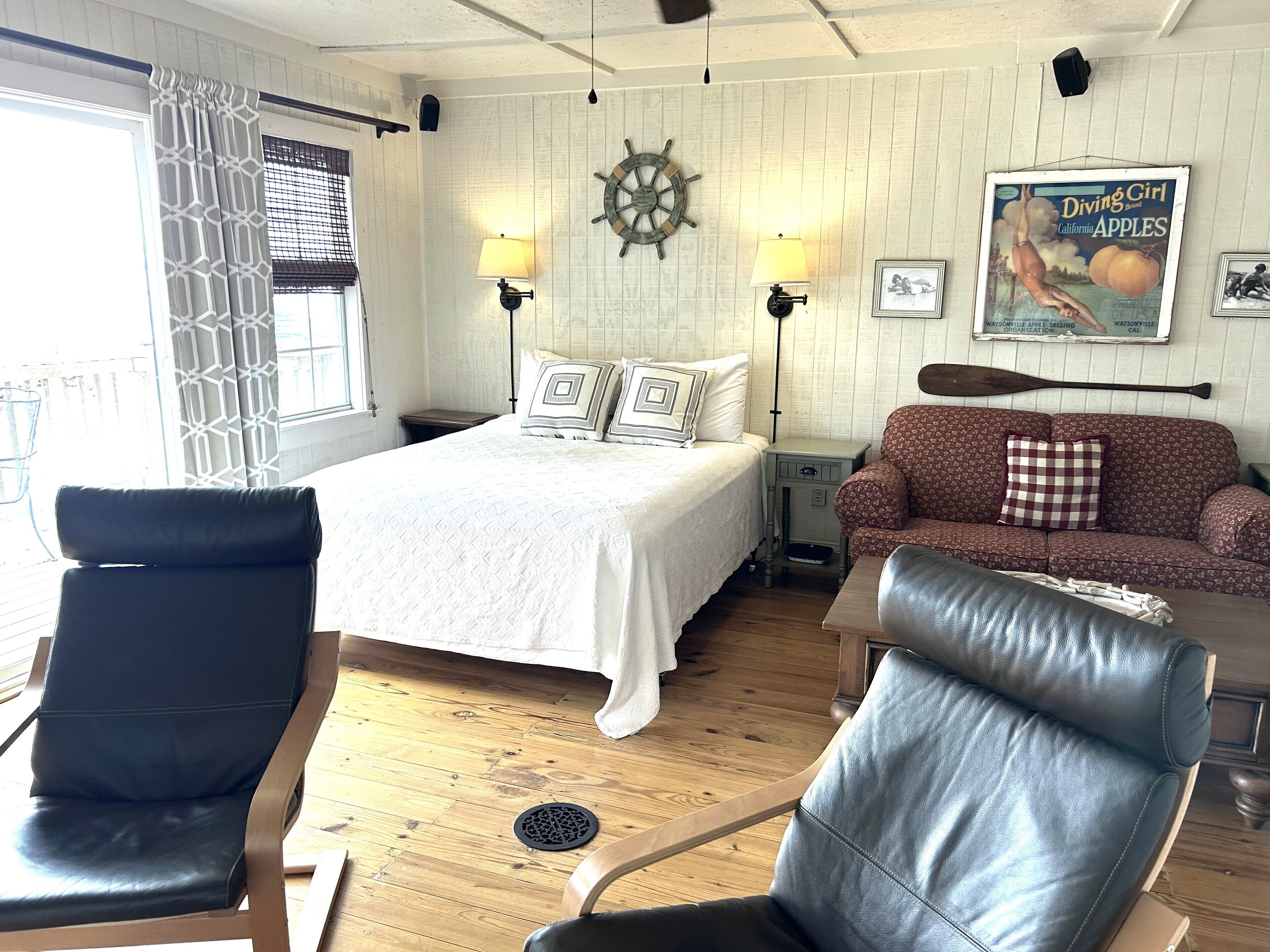 A room with a bed, two chairs, a sofa, a table, and a wall with a ship's wheel and a vintage magazine.