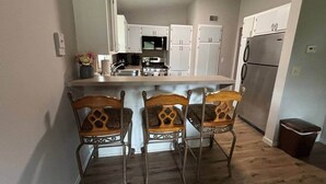 A kitchen with white cabinets, a stainless steel refrigerator, and a wooden dining table with three chairs. - Cedarwood Chateau (Cambridge)