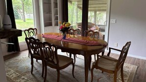 A dining room with a wooden table and chairs, a vase with flowers, and a view of the outdoors through large windows. - Cedarwood Chateau (Cambridge)