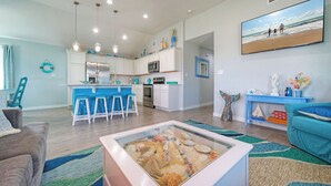 Uma cozinha moderna com armários brancos, uma ilha central com banquetas e uma área de jantar com sofá azul e mesa de centro com exposição de conchas.