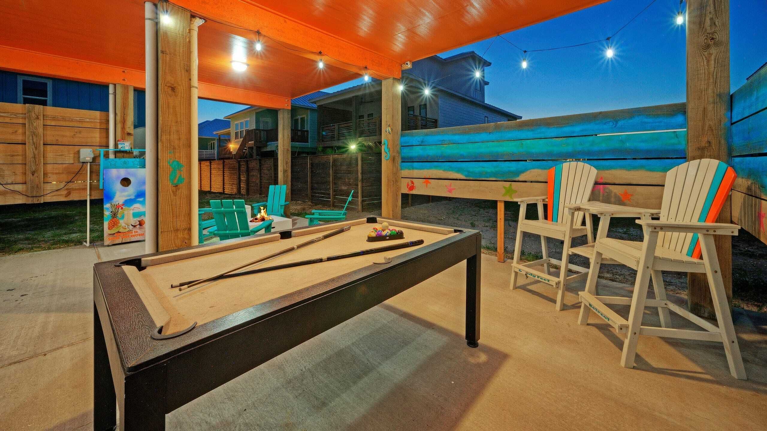 An outdoor pool table area with green chairs and a pool table, surrounded by wooden structures and string lights.