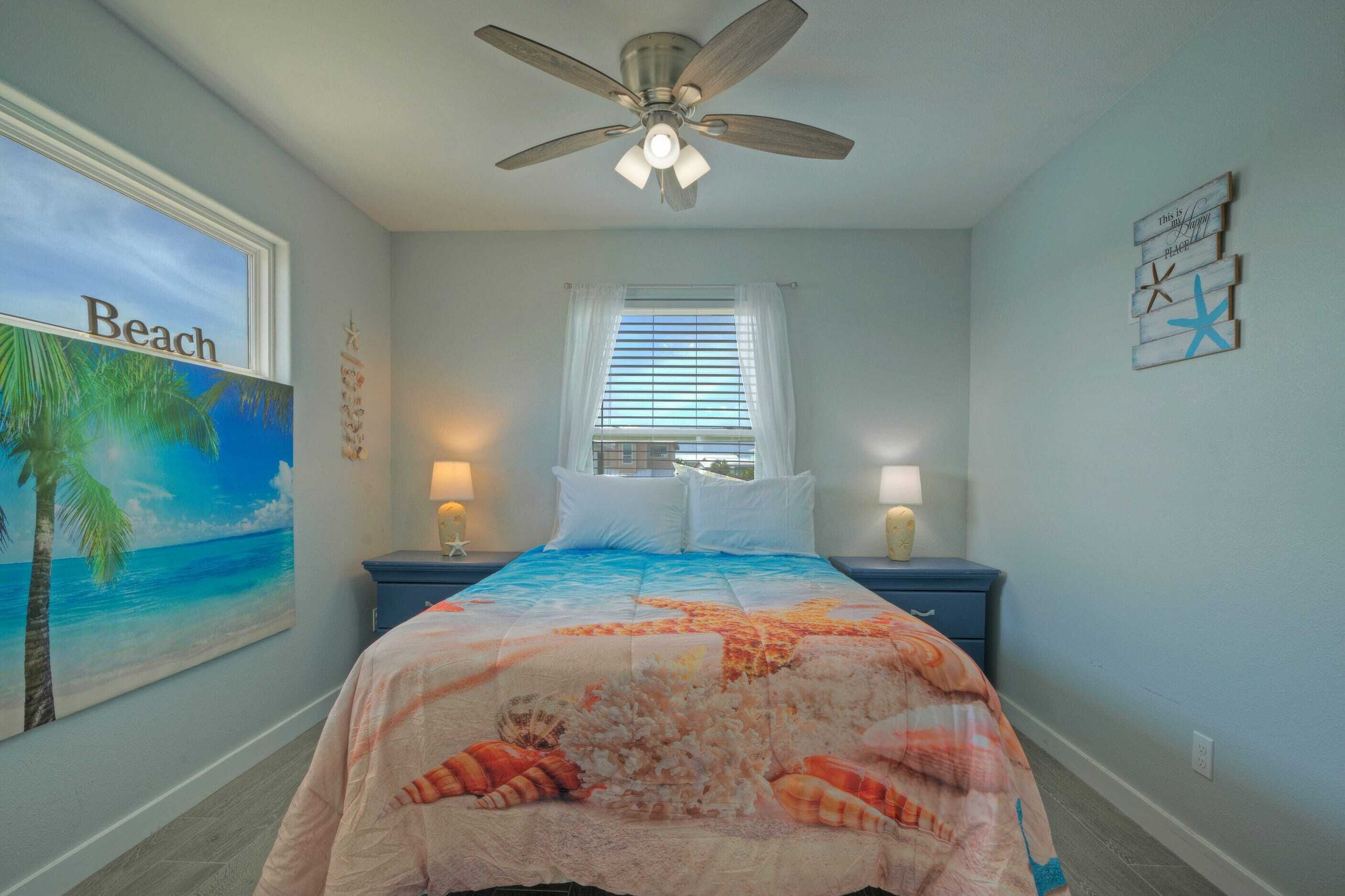 A bedroom with a bed, a ceiling fan, a window with blinds, a nightstand, and a wall with a painting and a starfish decoration.