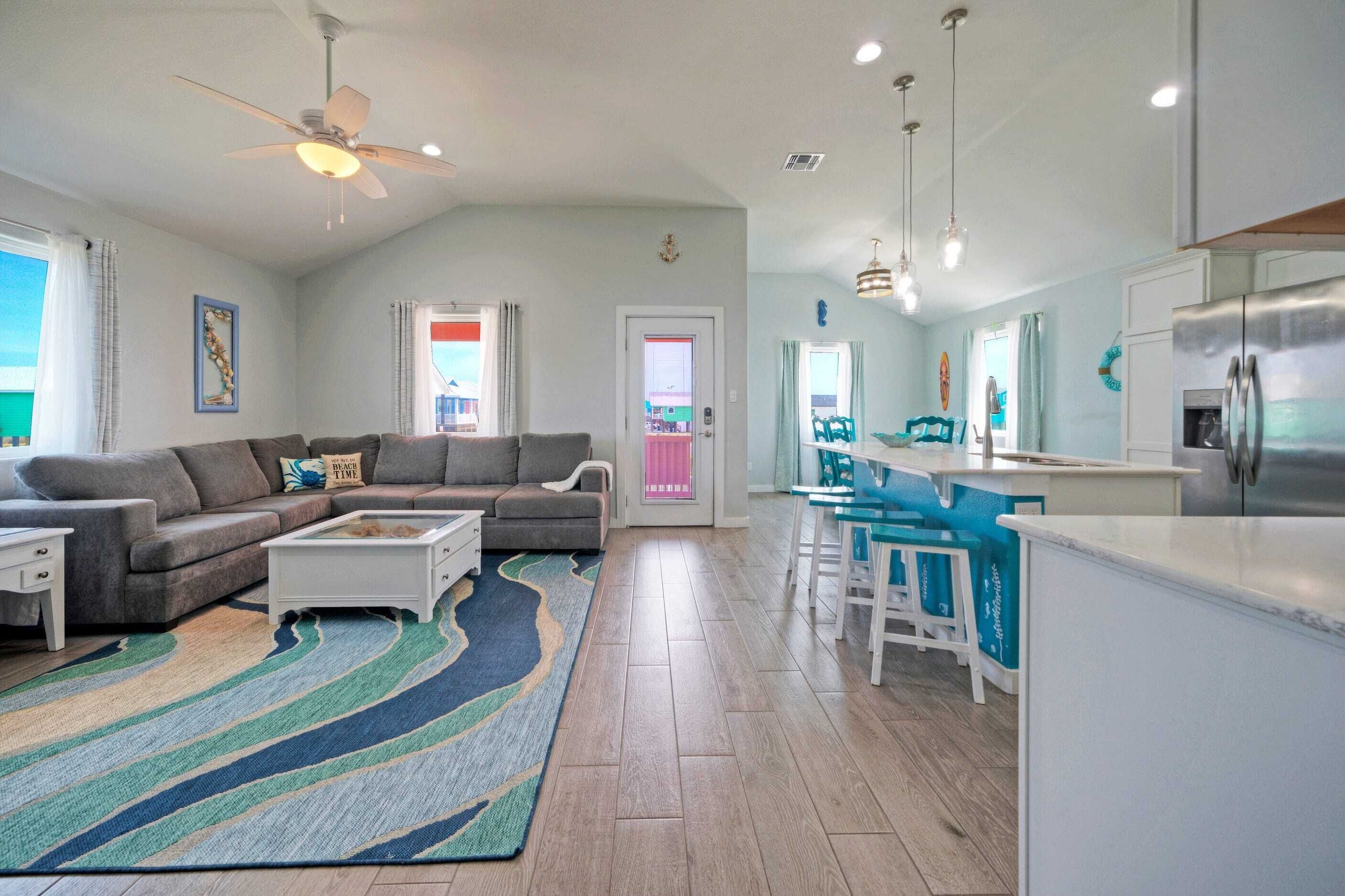 A modern living room with a sofa, coffee table, and a colorful rug. There is a dining area with bar stools and a kitchen with a refrigerator.