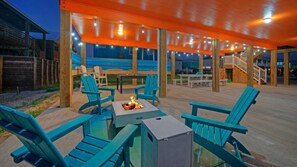 Een terras buiten met houten stoelen, een vuurkuil en een overdekte zithoek met lichtslingers.
