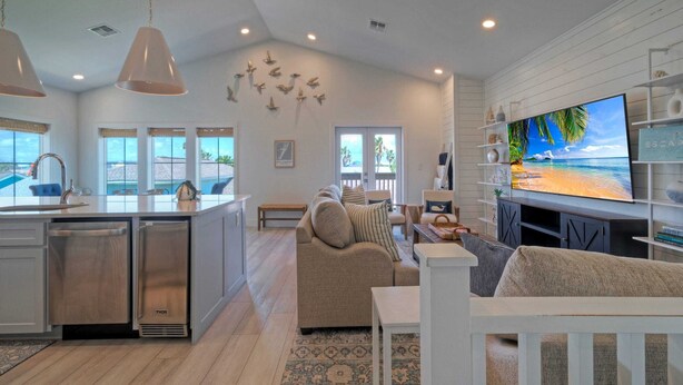 A modern kitchen with stainless steel appliances, a dining area with a white table and chairs, a living room with a beige sofa, and a large flat-screen TV displaying a beach scene. - Luxury 4bdr Beach Retreat w/ Private Pool Walk to Dining + Shops Shore Thang (Port Aransas)