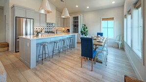 A modern kitchen with a central island, bar stools, and a dining area with a blue chair. - Luxury 4bdr Beach Retreat w/ Private Pool Walk to Dining + Shops Shore Thang (Port Aransas)