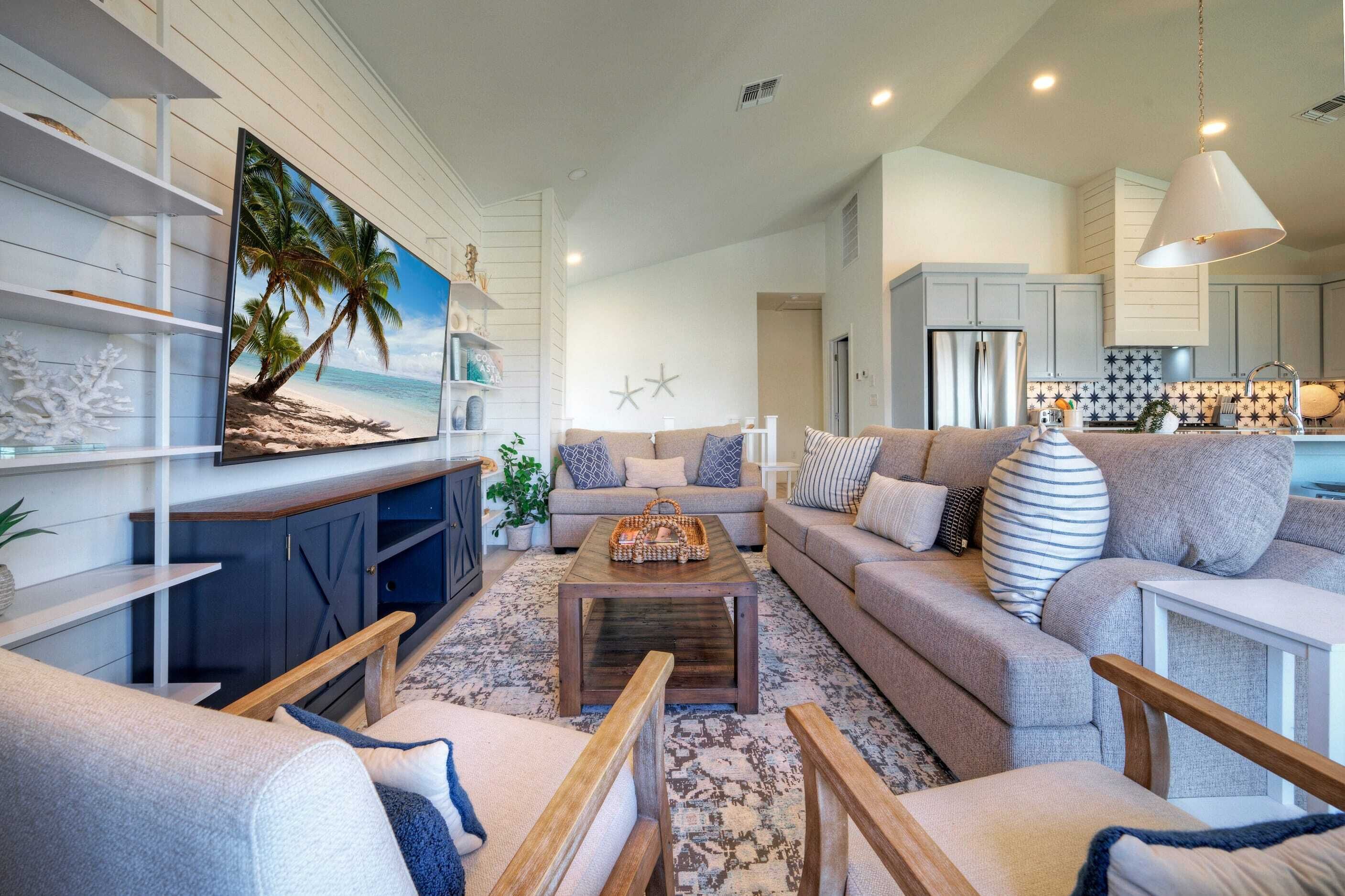 A modern living room with a large painting of a beach, a sofa set, a coffee table, and a kitchen area in the background.
