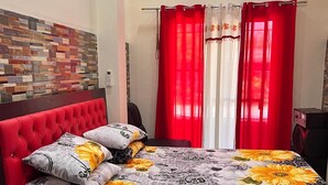 A bedroom with a red upholstered headboard, floral bedding, a brick-patterned wall, and red curtains.