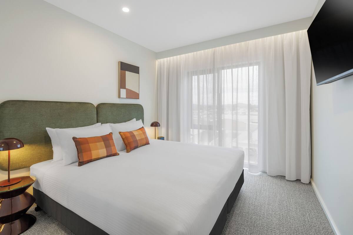 Ein Hotelzimmer mit einem großen Bett, einem grünen Kopfteil, zwei Nachttischen mit Lampen, einem Flachbildfernseher und einem großen Fenster mit durchsichtigen Vorhängen.