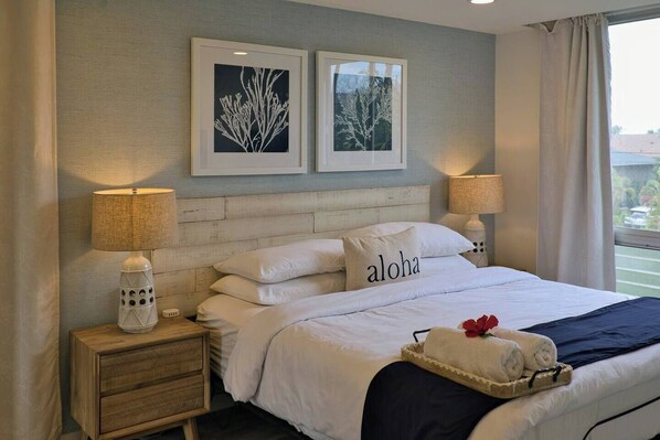 A neatly made bed with white linens, a pillow with the word "aloha," two framed botanical prints on the wall, and a window with curtains.