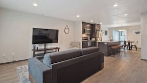 A modern living room with a large flat-screen TV, a grey sofa, a small kitchen area with dark cabinets, and a dining space with a bar. - Your stay will include the entire Unit A to yourself (Spokane)
