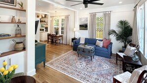 A spacious living room with a blue sofa, a coffee table, and a patterned rug. There is a dining area with a wooden table and chairs, and a kitchen in the background. - Riverwalk Mill Cottage - Southern Charm Stay (Columbus)