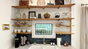 A kitchen with a sink, shelves with various items, a window with a view, and a coffee machine. - Riverwalk Mill Cottage - Southern Charm Stay (Columbus)