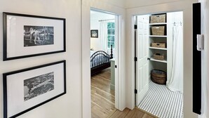 A hallway with framed black and white photographs on the wall, a bed with a patterned bedspread, and a basket-filled shelf. - Riverwalk Mill Cottage - Southern Charm Stay (Columbus)