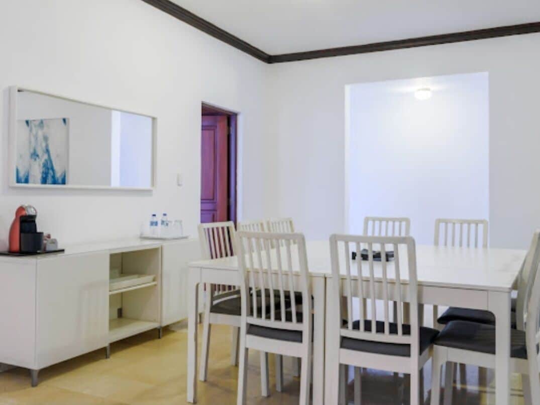 Un comedor con una mesa y sillas blancas, un armario blanco con una cafetera y un cuadro en la pared.