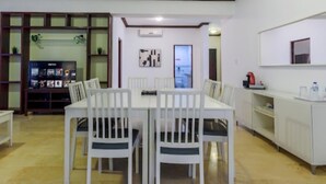 Un comedor con una mesa y sillas blancas, un televisor sobre un mueble bajo y una cocina equipada con refrigerador y microondas.