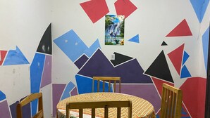 Ein Essbereich mit einem runden Tisch, bedeckt mit einer blumigen Tischdecke, umgeben von Holzstühlen. Die Wände sind mit farbenfrohen geometrischen Mustern und einem gerahmten Bild von einem Wasserfall dekoriert.