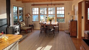 Interior acogedor de una cabaña con una mesa de comedor de madera, sillas, chimenea y grandes ventanales que ofrecen vistas al exterior.
