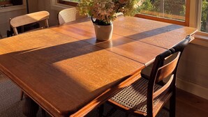 Un comedor con mesa y sillas de madera, una planta en maceta con flores blancas y grandes ventanas con vistas a un cuerpo de agua y árboles.