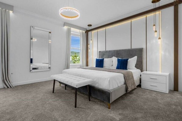 A modern bedroom with a large bed, a bench, a nightstand, a mirror, and a view of greenery outside.