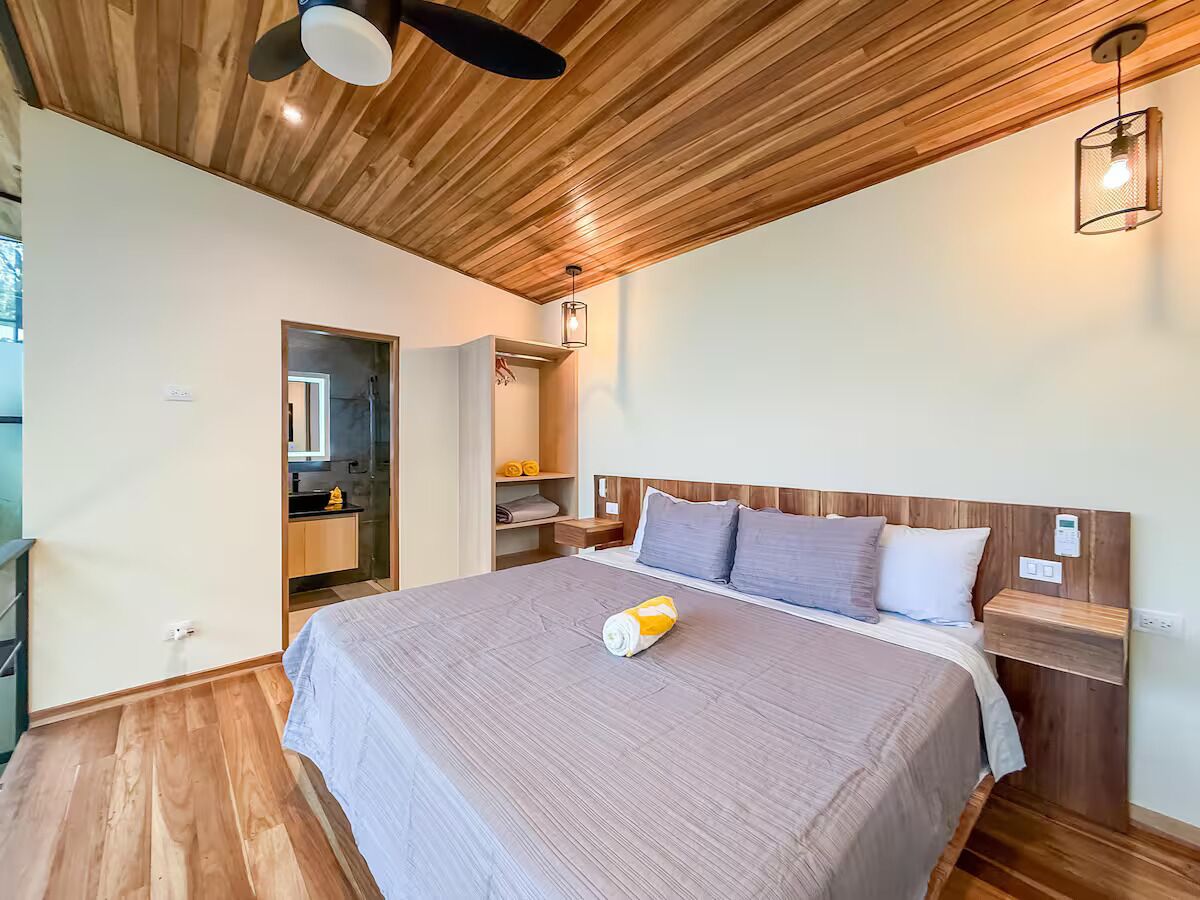 Quarto com teto de madeira, cama com colcha cinza, mesa de cabeceira e criado-mudo.