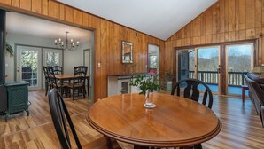 Un comedor con mesa y sillas de madera, una araña y vistas al exterior a través de ventanas amplias.