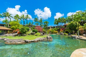 Piscina cristalina con rocas y palmeras, rodeada de vegetación exuberante y un cielo azul despejado.
