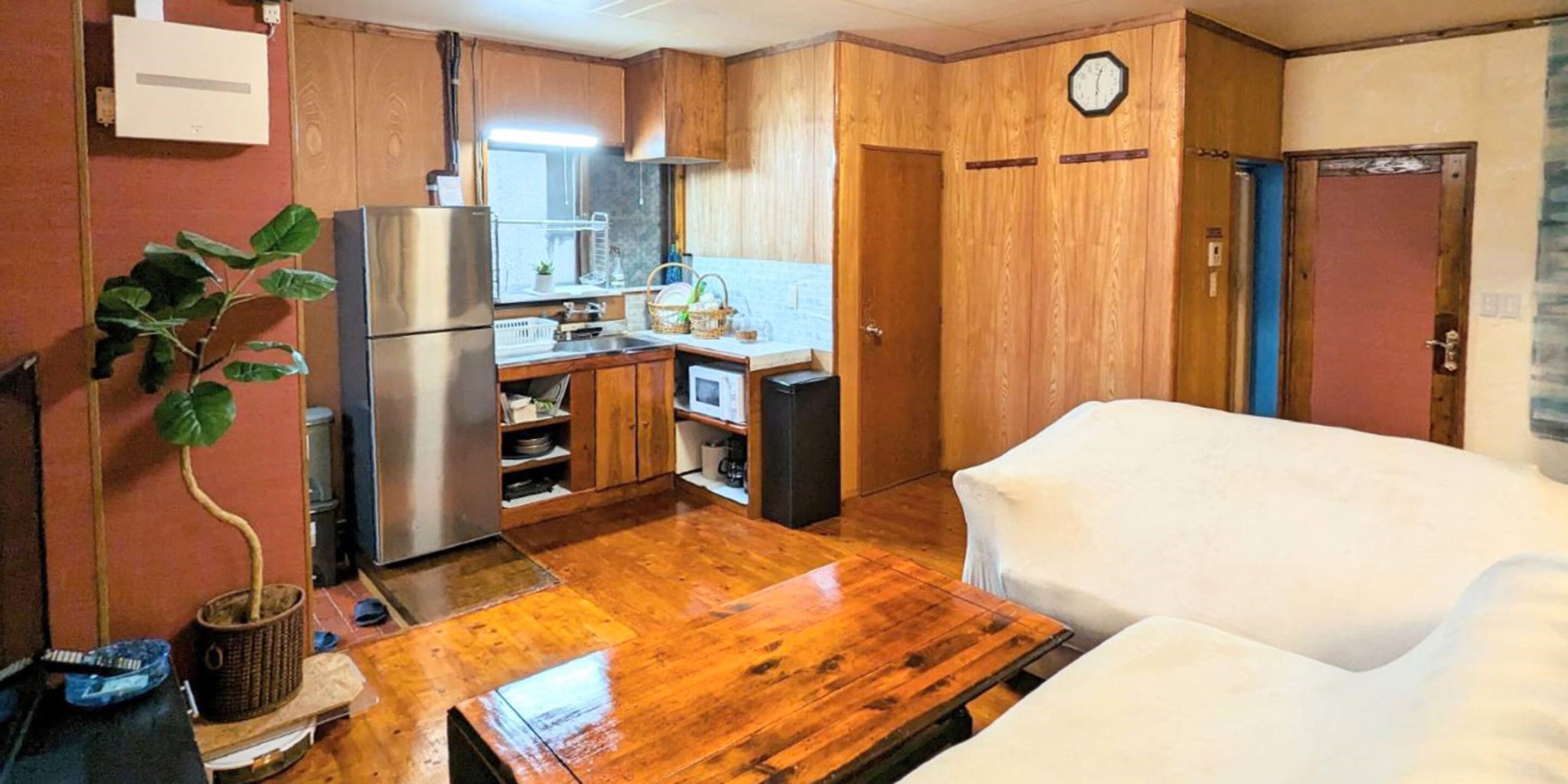 A compact kitchen with wooden cabinets, a refrigerator, a microwave, and a sink. There is a clock on the wall and a door to the right.