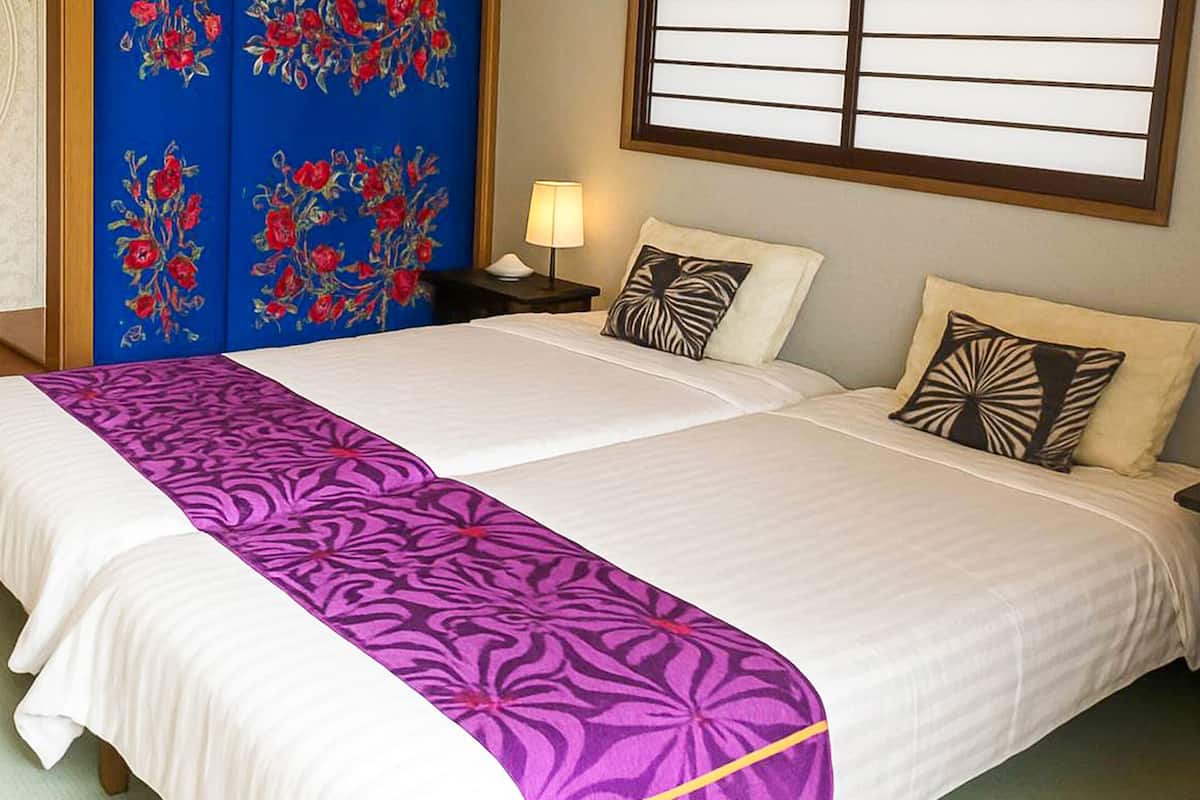 Quarto de hotel com cama, mesas de cabeceira, luminária e janela com persianas.